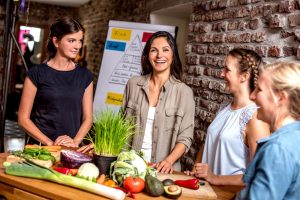 Vier Personen in einem Workshop der Weiterbildung Vegane Ernährungsberatung stehen an einem Tisch mit Gemüse neben Flipchart