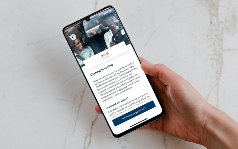 Smartphone in der Hand. Auf dem Bildschirm zu sehen ist ein Beitrag in der Earnest App mit der Überschrift "Sharing is caring" zum Thema Fahrgemeinschaften